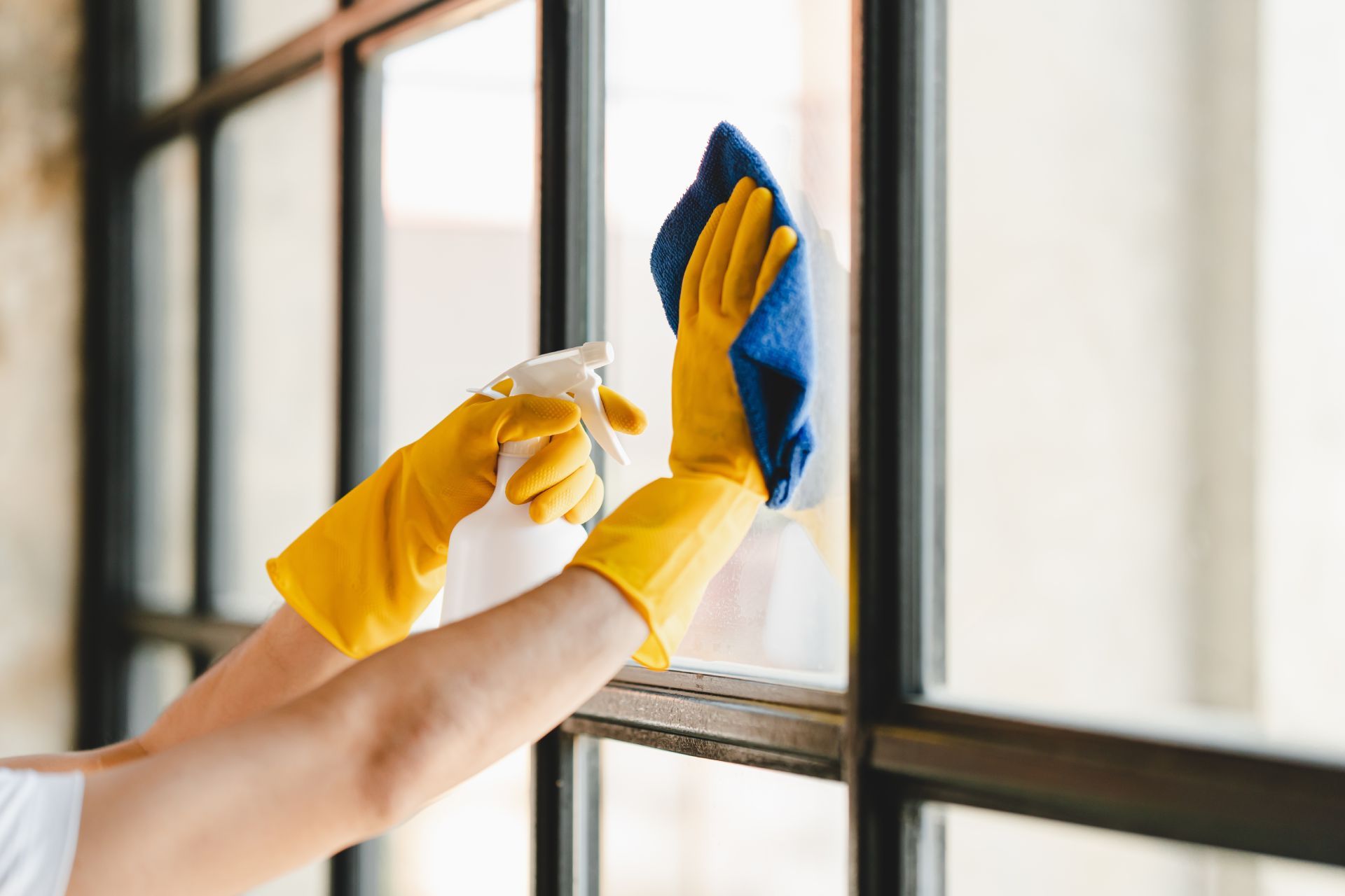 Person wearing yellow gloves spraying and wiping a window with a blue cloth.