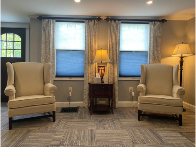 Two beige wingback chairs face each other in a room with a patterned carpet and two windows with sheer curtains.