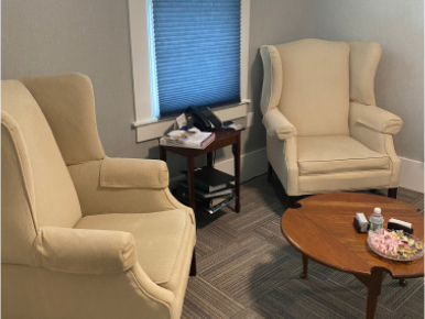 Two cream wingback chairs face each other in a room with a small table, a round table, and a window.