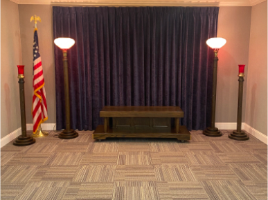 Funeral home interior: dark blue curtain backdrop, wooden table, lamps, US flag, and red candle holders.