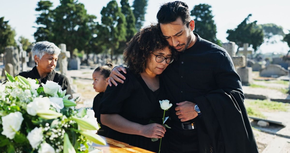 People at a funeral, hugging at a gravesite. Woman holding a white rose, other mourners in background.
