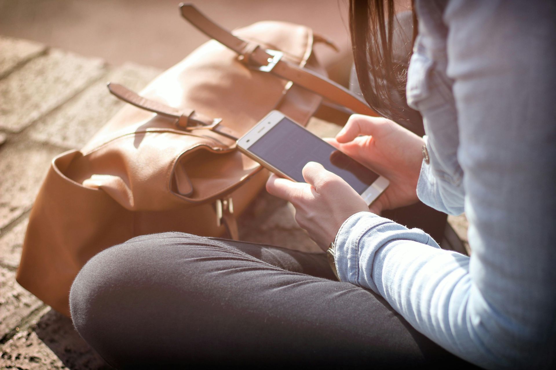 Woman sitting, using smartphone, with a tan bag beside her.