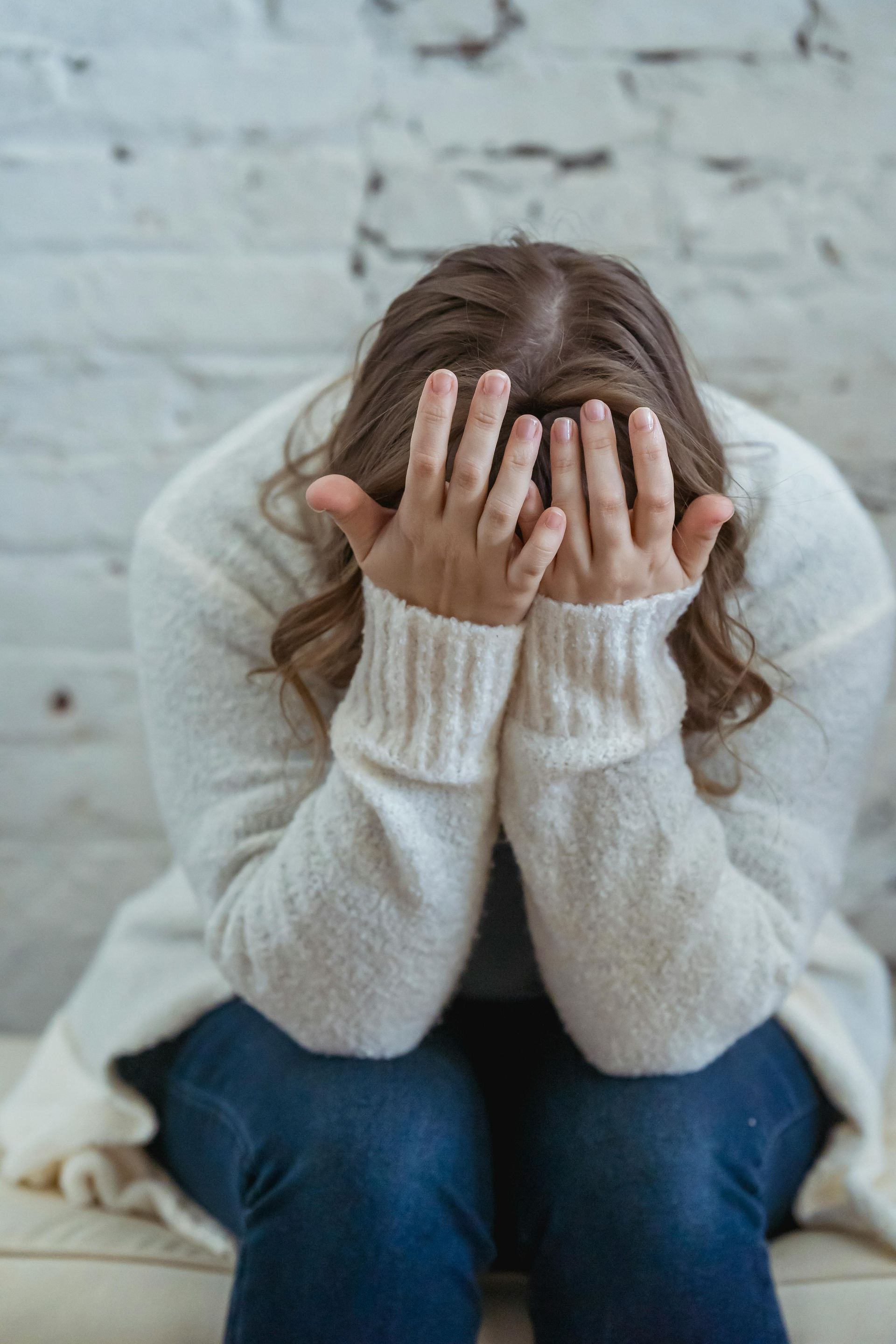 Woman sitting, head in hands, appears distressed. Wearing blue jeans and a white sweater. Against a white brick wall.