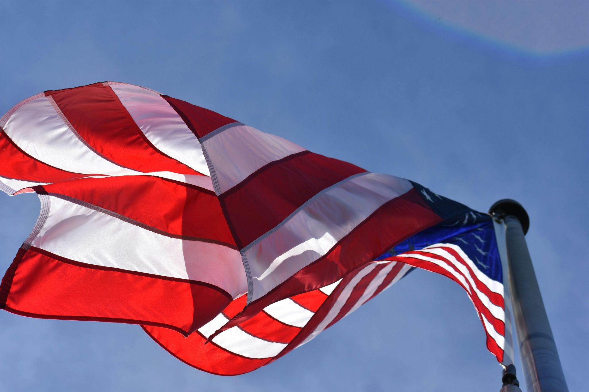 American flag waving in the wind against a blue sky. Stripes of red and white, stars in a blue field.