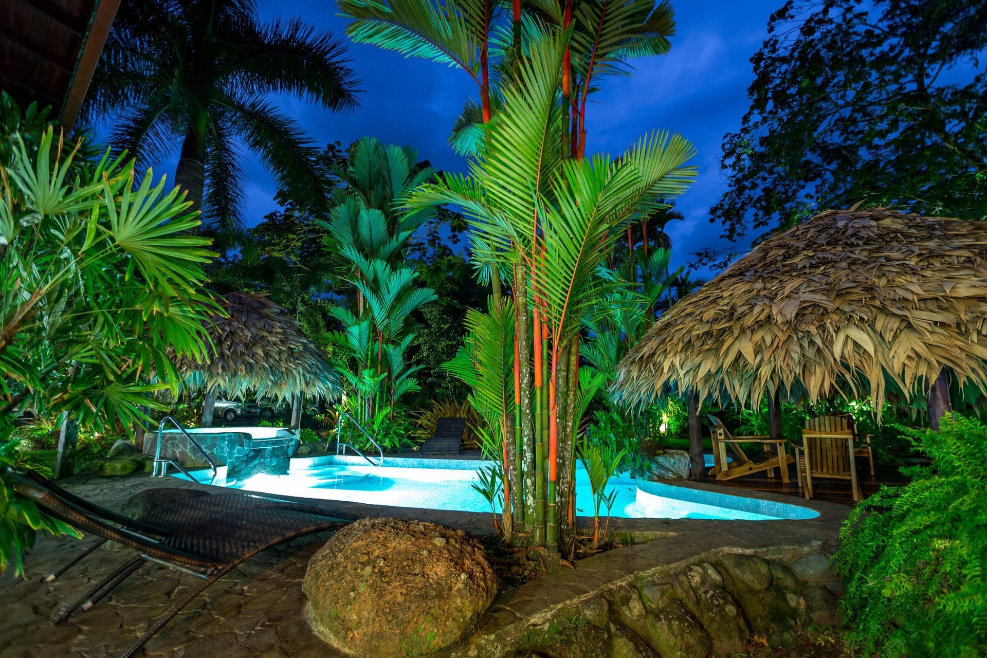A swimming pool is surrounded by palm trees and thatched umbrellas at night.