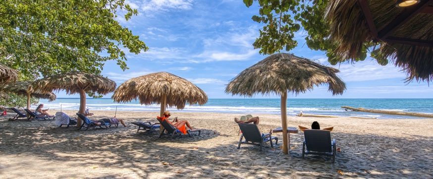A group of people are sitting under thatched umbrellas on a beach.