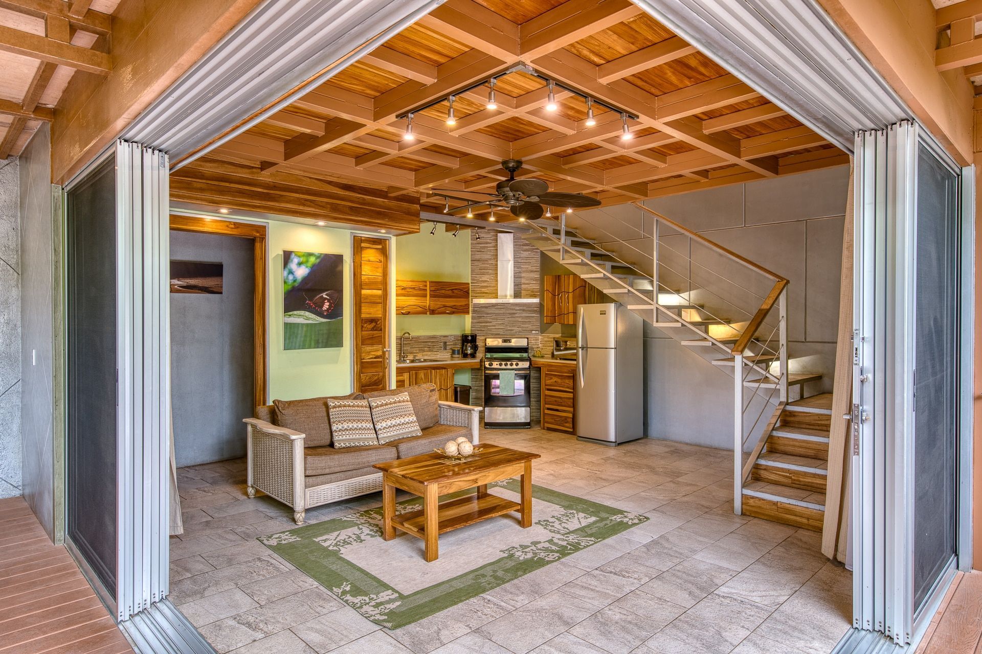 A bright, open-plan interior with a living room, kitchenette, and staircase, seen through a wide glass sliding door.