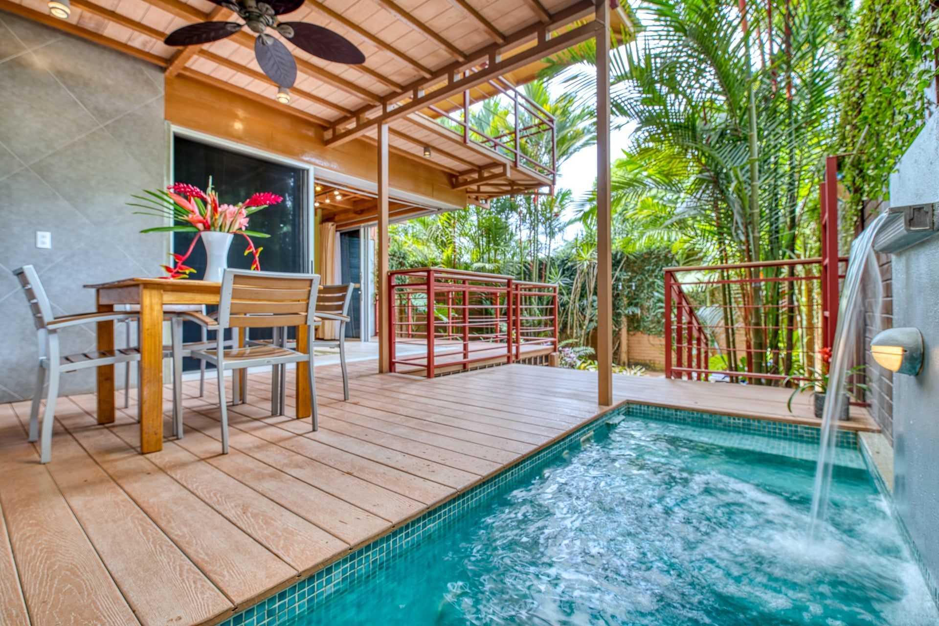 A wooden deck featuring a dining table and chairs beside a small, turquoise swimming pool with a stone water feature.