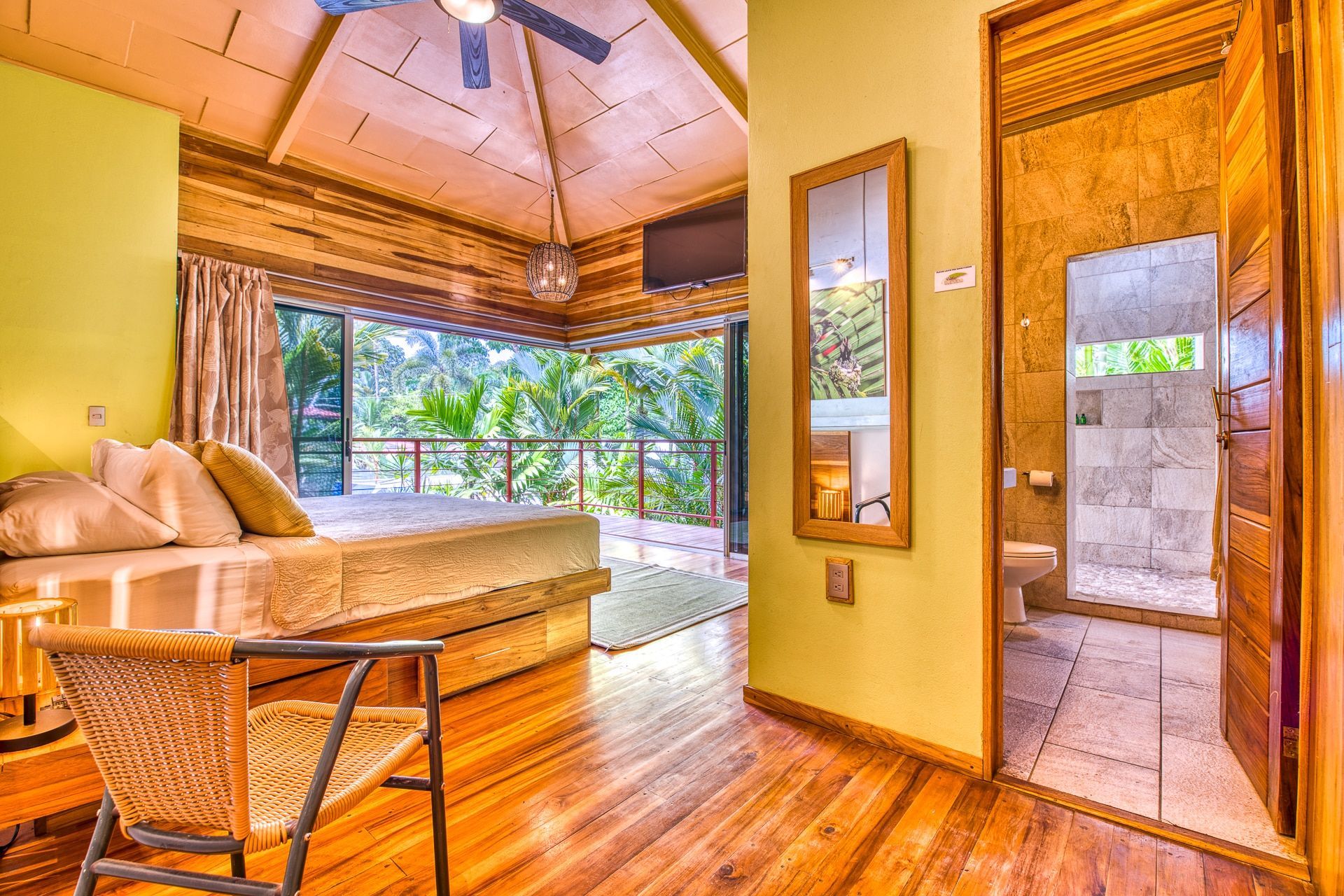 A sunlit bedroom with a wooden floor, a bed with light-colored pillows, a woven chair, and an open door to a bathroom.