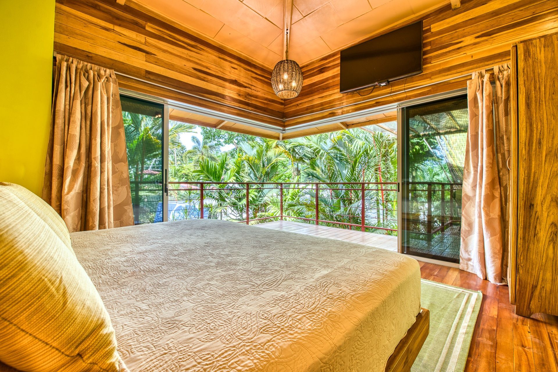 A bedroom with wood-paneled walls and ceilings, featuring a large bed and glass doors overlooking a tropical garden.