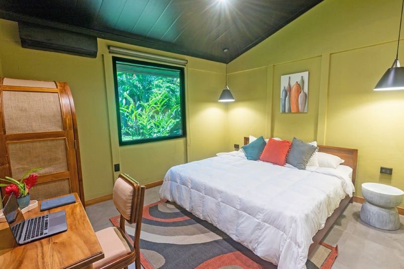 A modern, earth-toned bedroom with a white-bedded king bed, a wooden desk with a laptop, and a view of lush greenery.