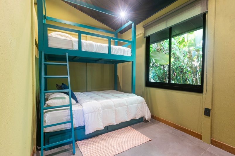A teal bunk bed with white bedding in a room with light green walls, a black ceiling, and a window overlooking greenery.