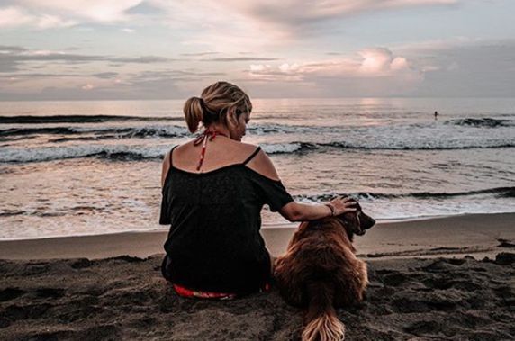 A woman and her dog are sitting on the beach looking at the ocean.