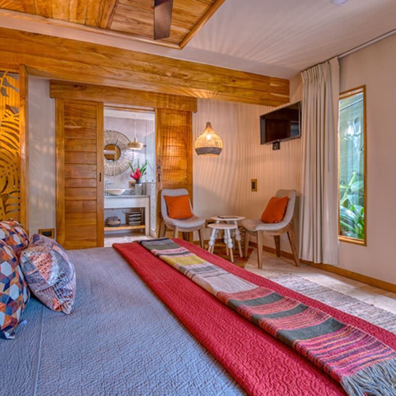 A bedroom with a large bed, colorful patterned pillows, a red runner, two chairs, a small table, and an open bathroom.