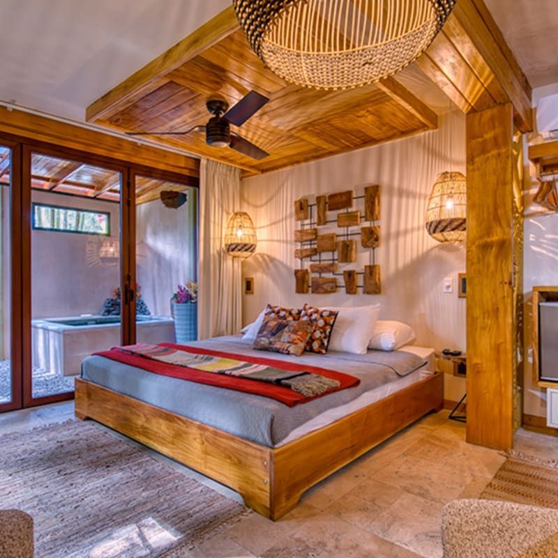 A bedroom with wooden ceilings, a ceiling fan, a woven pendant light, and a bed with gray bedding and a red throw blanket.