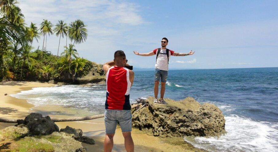A photographer in a red and white vest takes a picture of a person standing on a rock with arms open by the ocean.