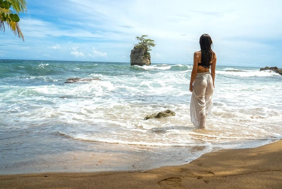 A woman is standing in the water on a beach looking at the ocean.