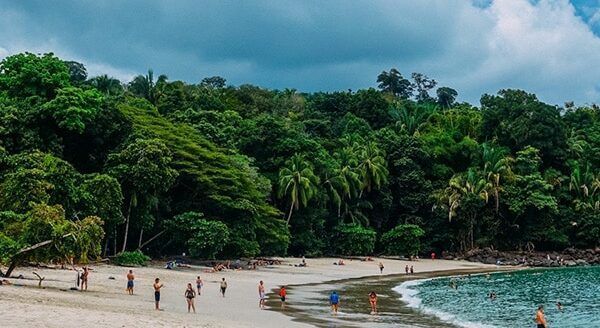 People relax on a sandy beach fronted by dense, green forest under a cloudy sky.