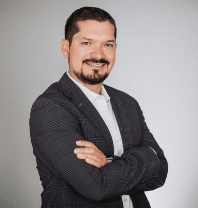 A man with a beard is wearing a suit and smiling with his arms crossed.