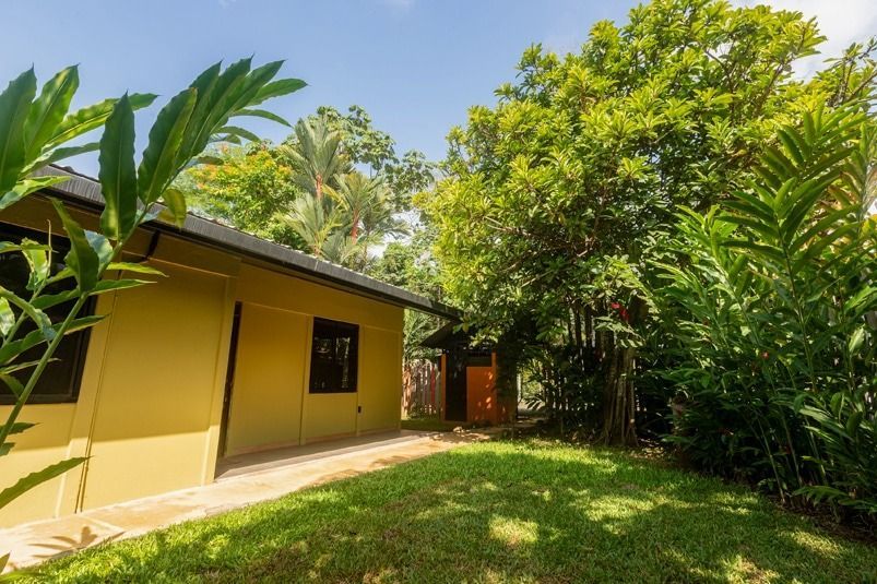 A yellow building with a dark roof surrounded by lush green trees and plants under a blue sky.