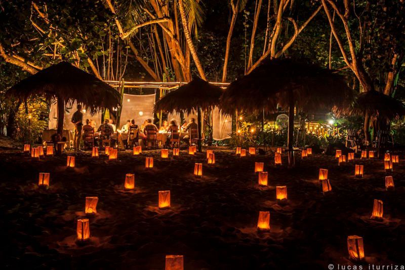 A group of people are sitting under thatched umbrellas surrounded by candles.
