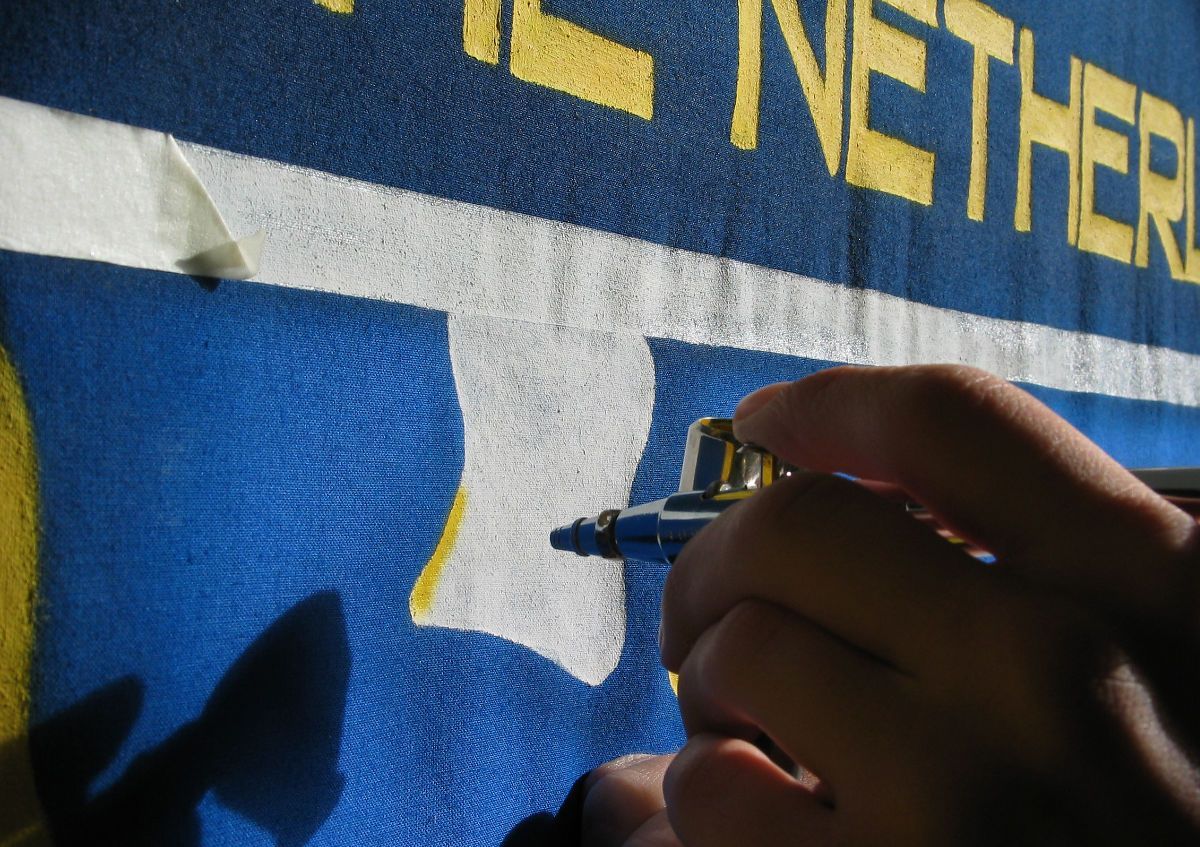 Een hand kleurt met een blauwe stift een gele vorm op blauwe stof. Het woord