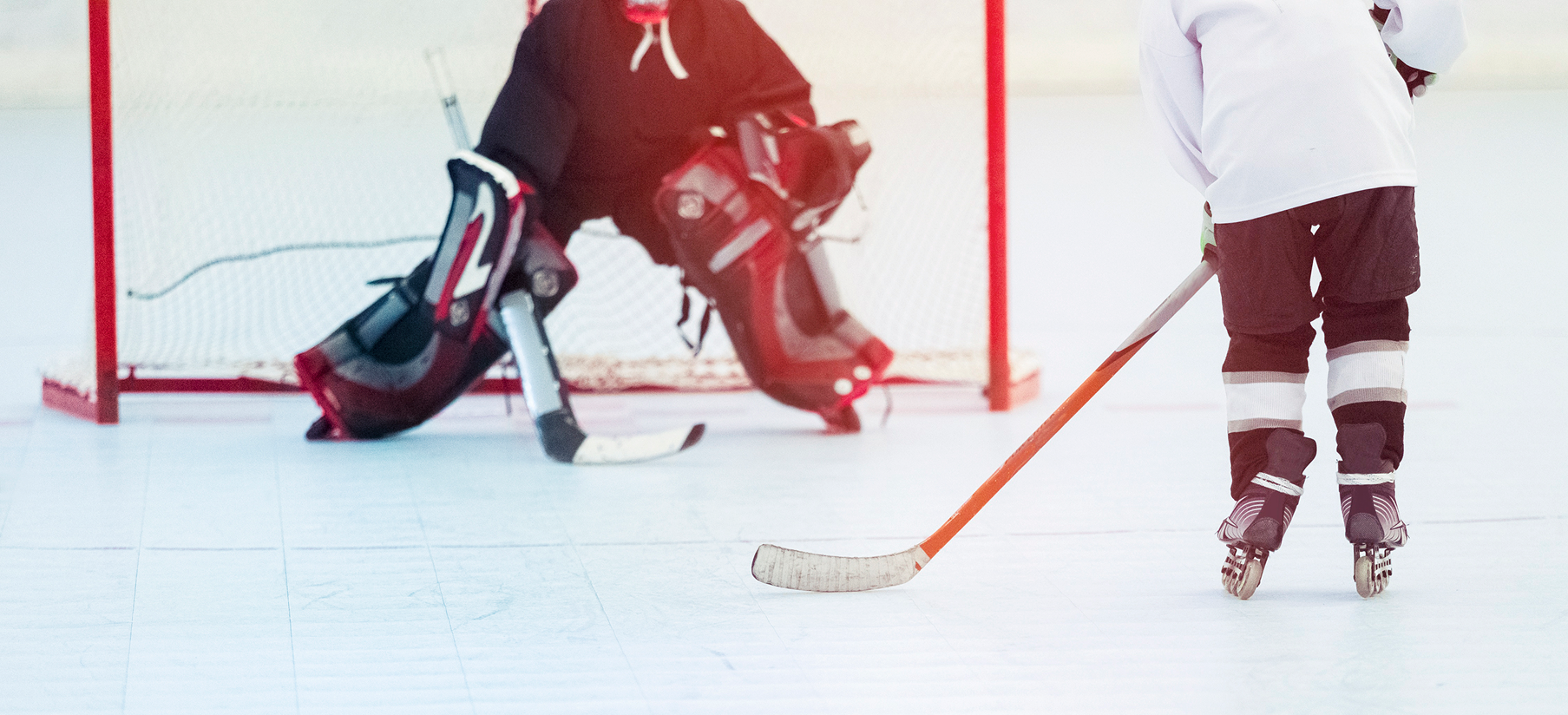Hockey game in progress. A goalie in the net faces a player with a hockey stick, ready to shoot the puck.