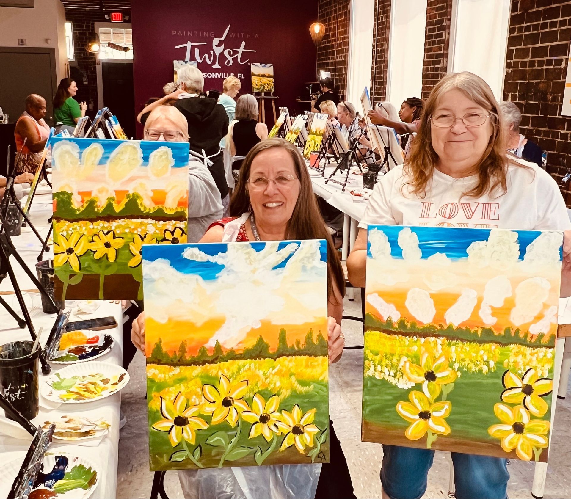 Two women are holding up paintings of a field of sunflowers.