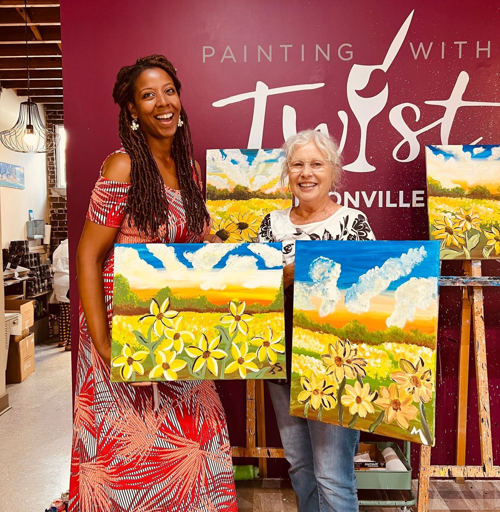Two women are holding paintings in front of a sign that says painting with twist