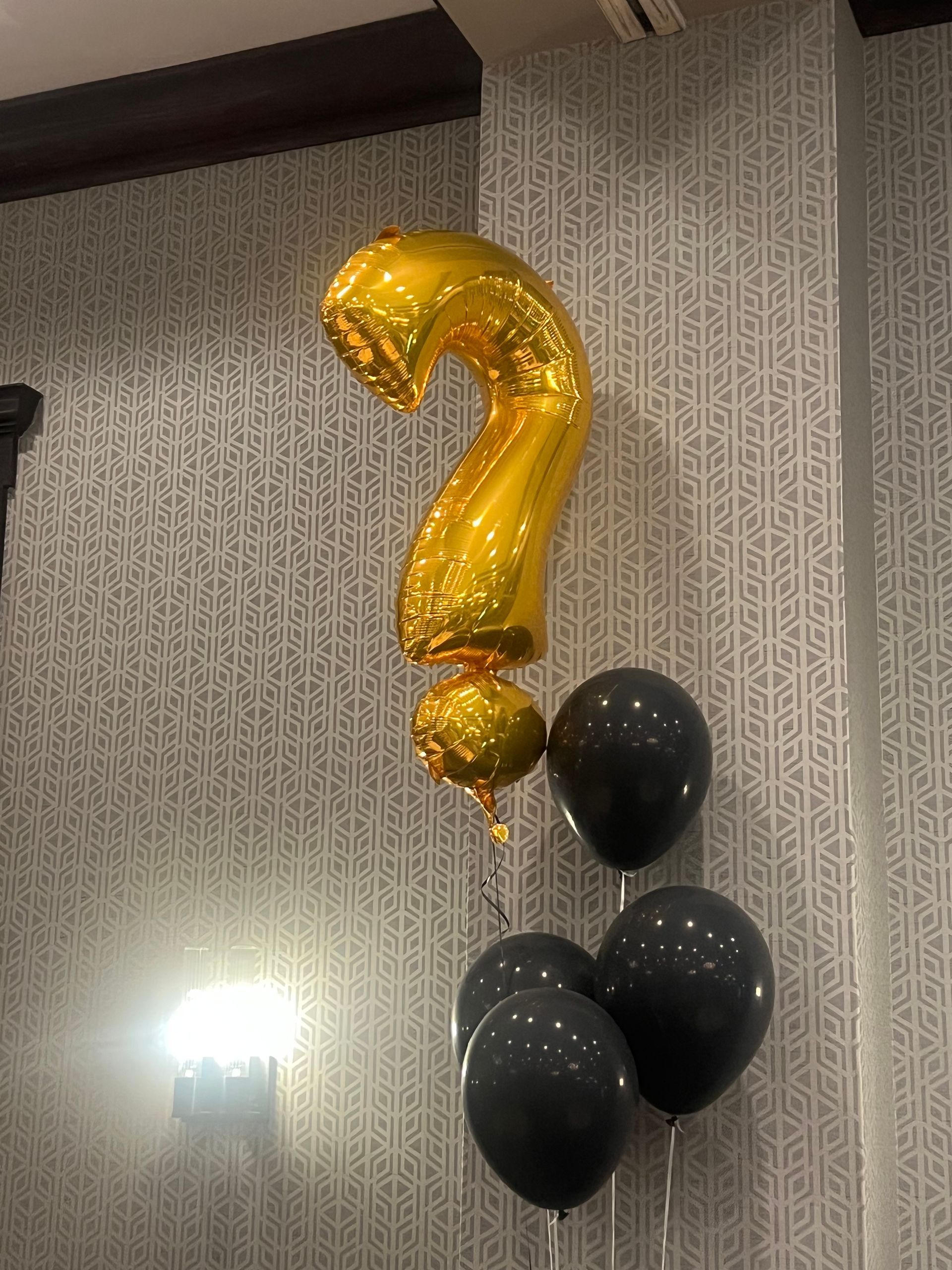 Gold question mark balloon with black balloons on a patterned wall.