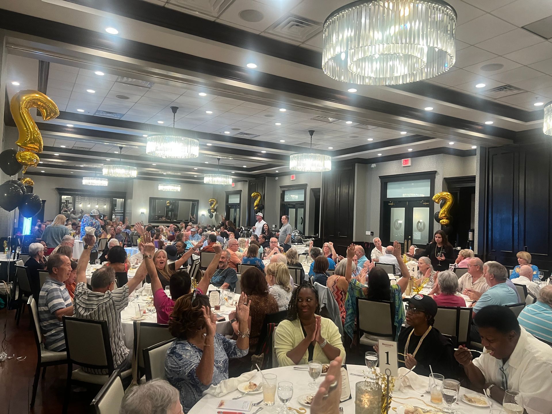 Large gathering in a banquet hall, many people seated at tables, some raising hands. Gold balloons present.