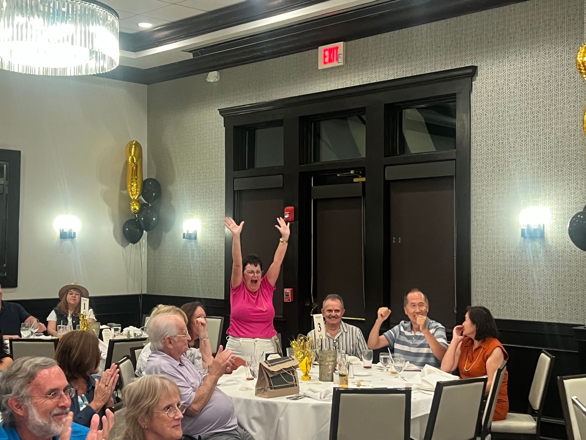 People celebrating at a table in a decorated room; a woman raises her arms in excitement.