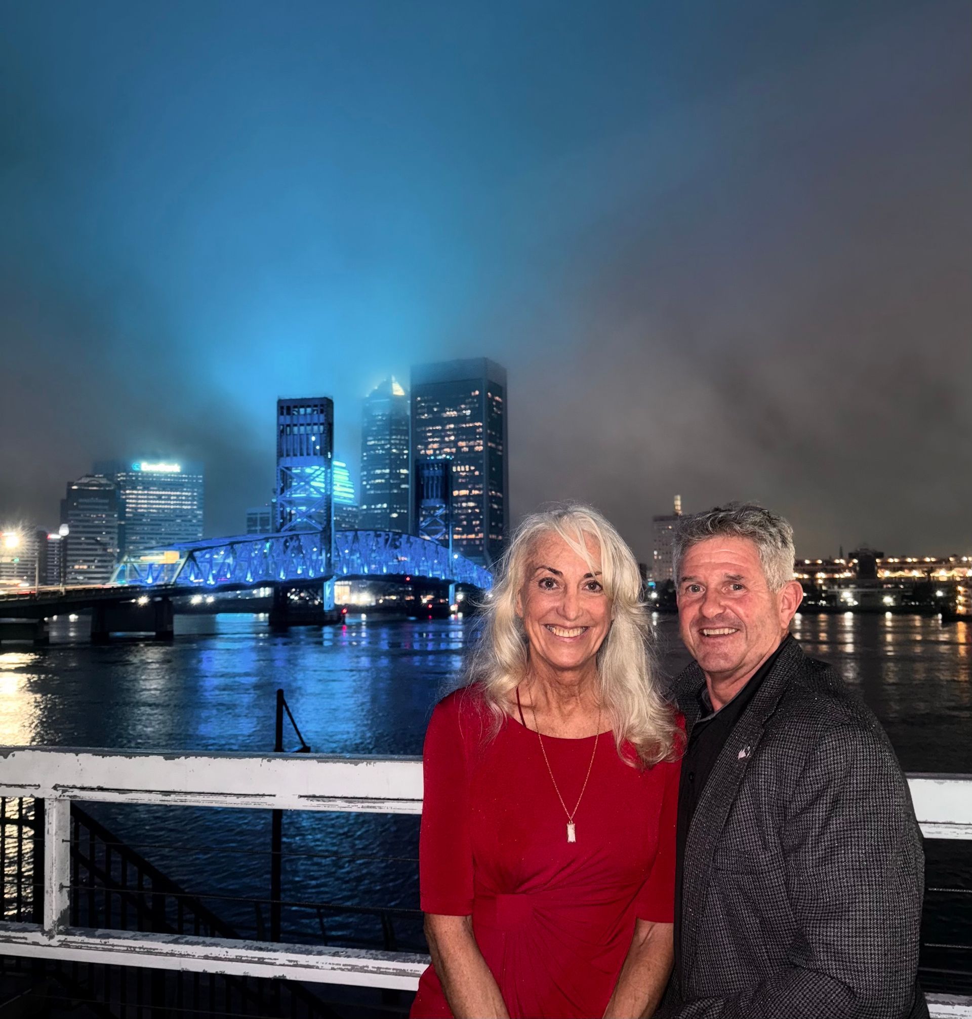 A man and a woman are posing for a picture in front of a bridge over a body of water.
