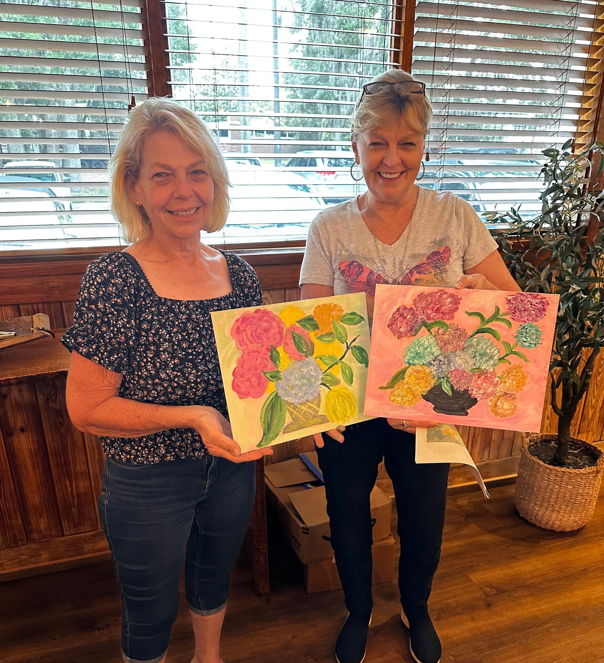 Two women are standing next to each other holding paintings of flowers.