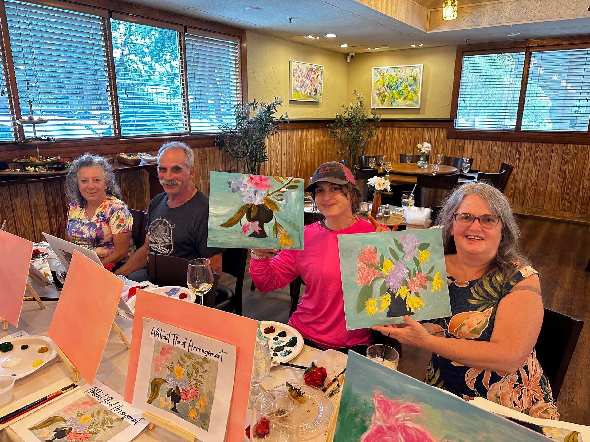 A group of people are sitting at a table holding up paintings.