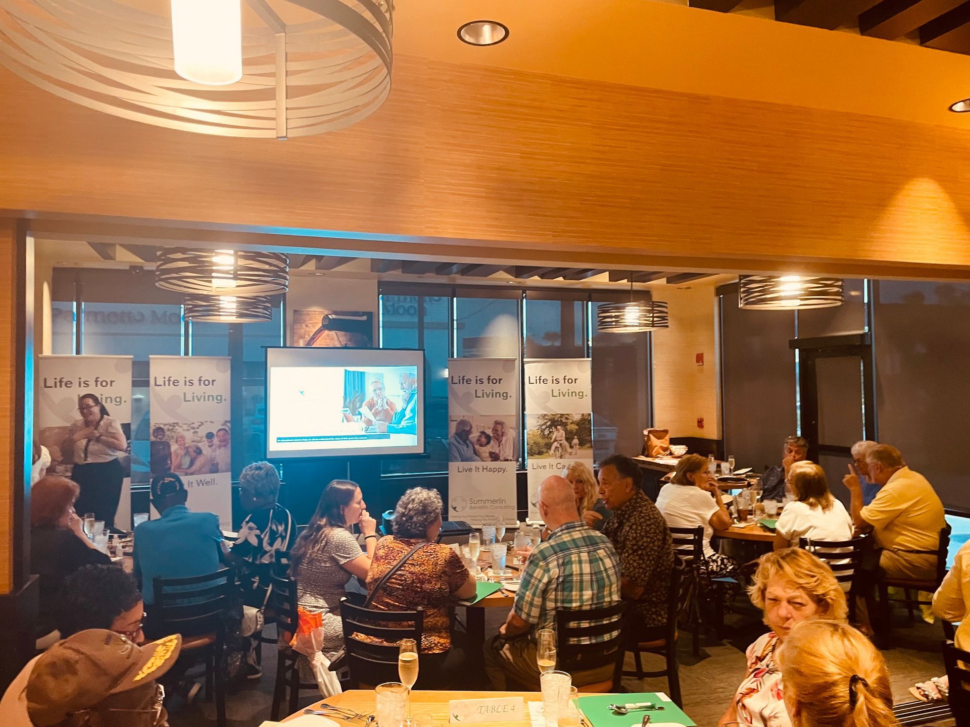 A group of people are sitting at tables in a restaurant watching a presentation