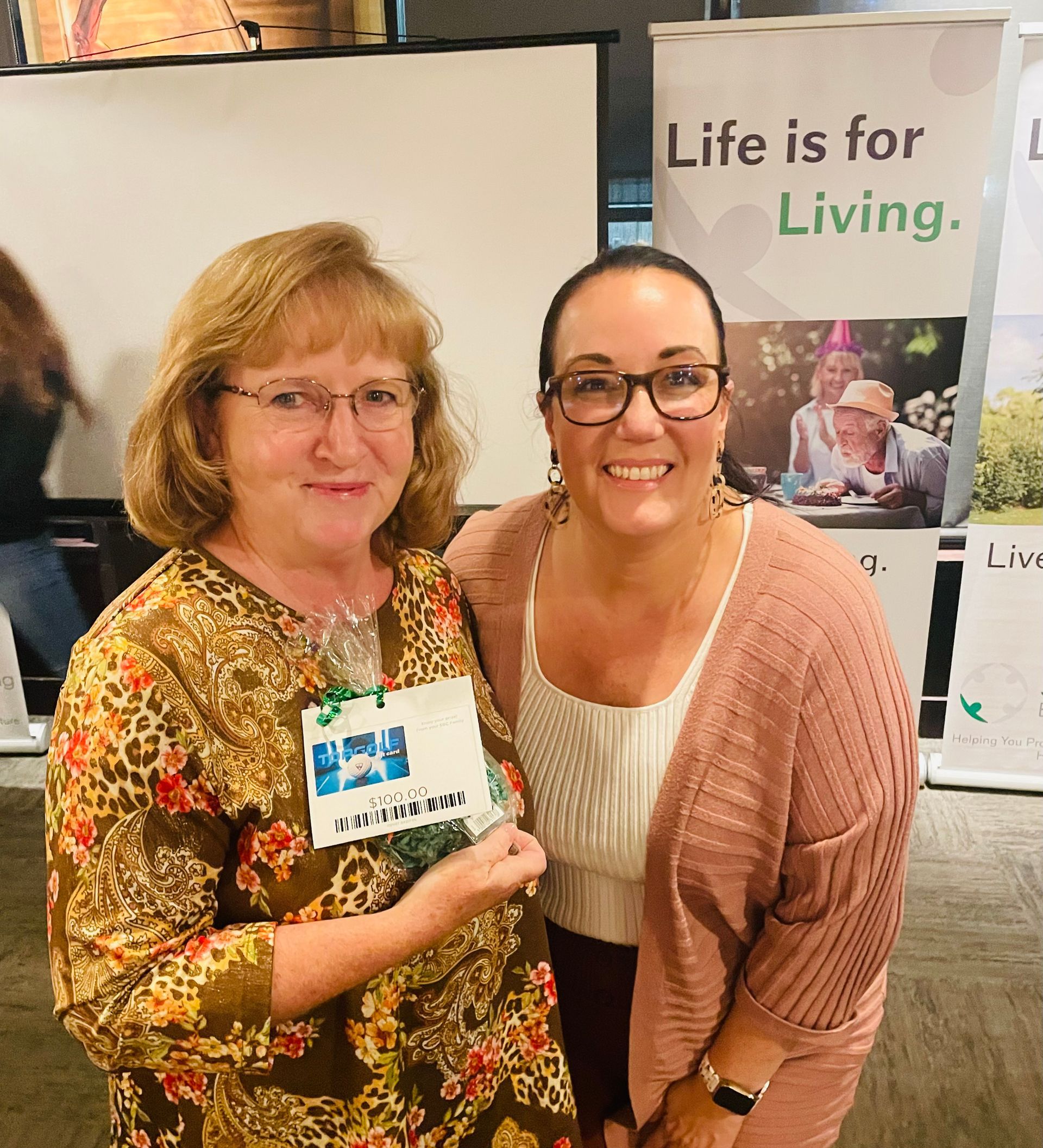 Two women are posing for a picture in front of a sign that says life is for living