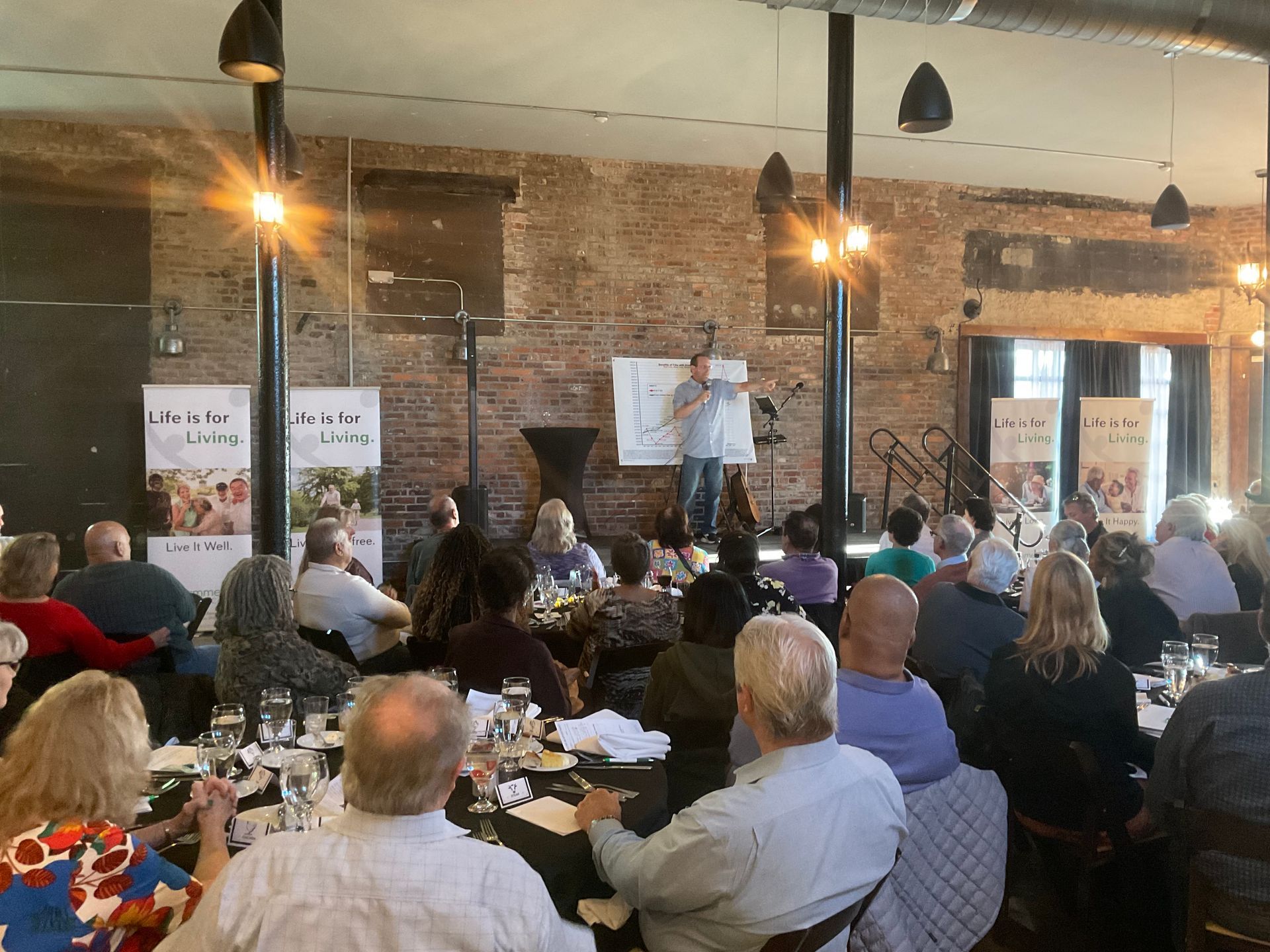 A man is giving a presentation to a large group of people sitting at tables.