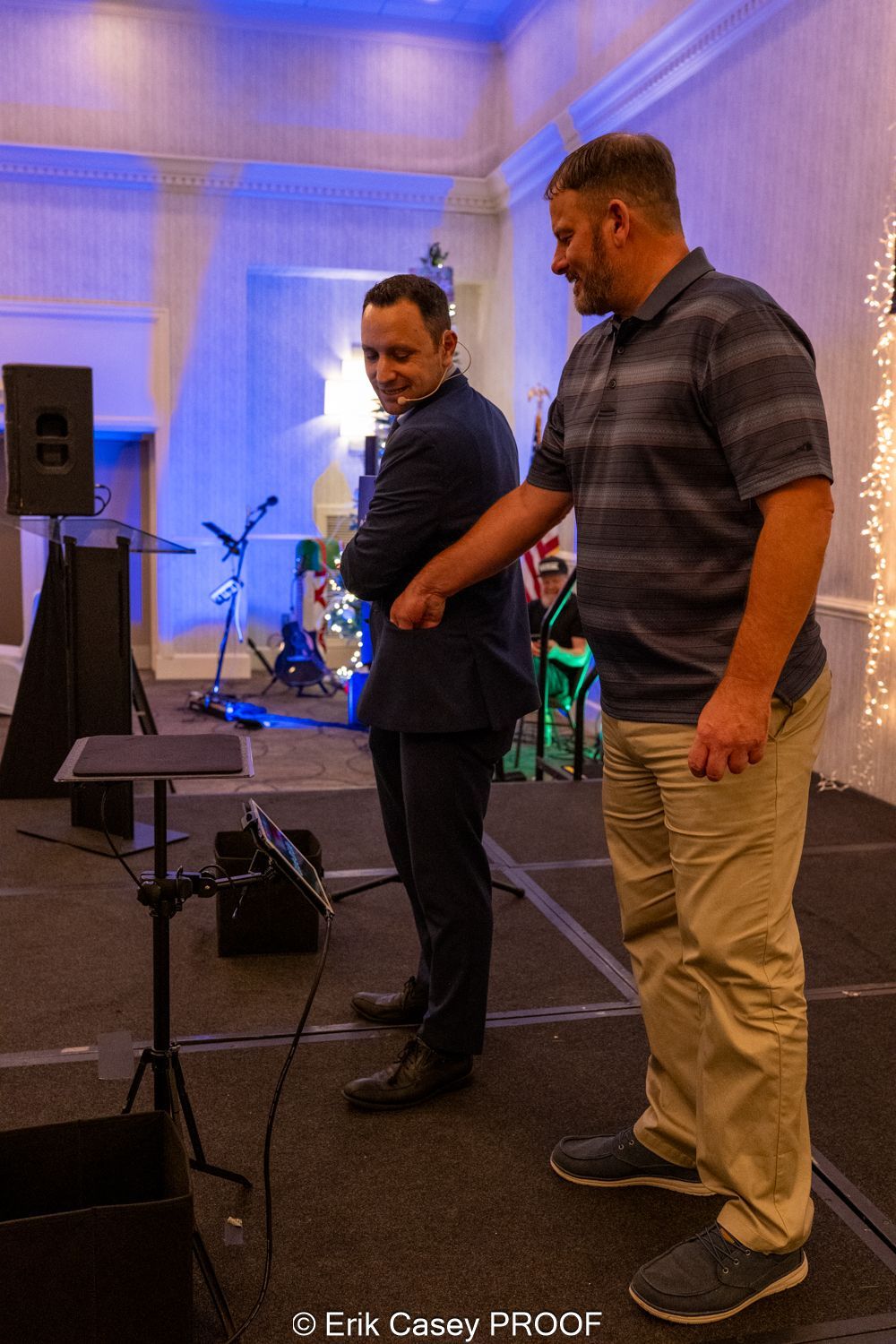 Two men are standing next to each other on a stage in a room.