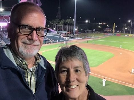A man and a woman are posing for a picture in front of a baseball field.