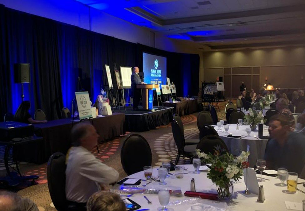 A man is giving a speech at a conference in a large room with tables and chairs.