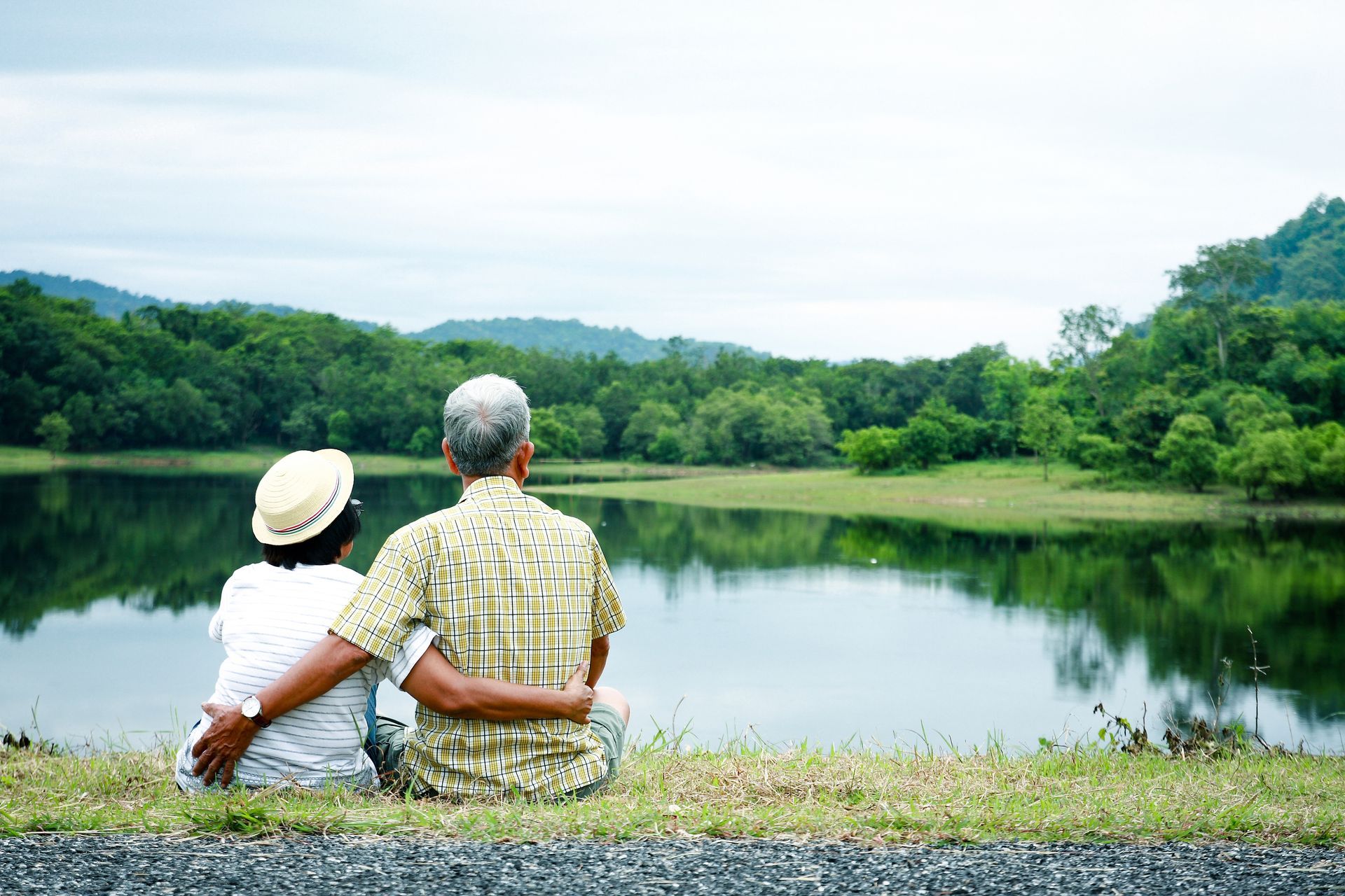 Man and woman with arms around each other's backs sitting in the grass overlooking a small lake. 