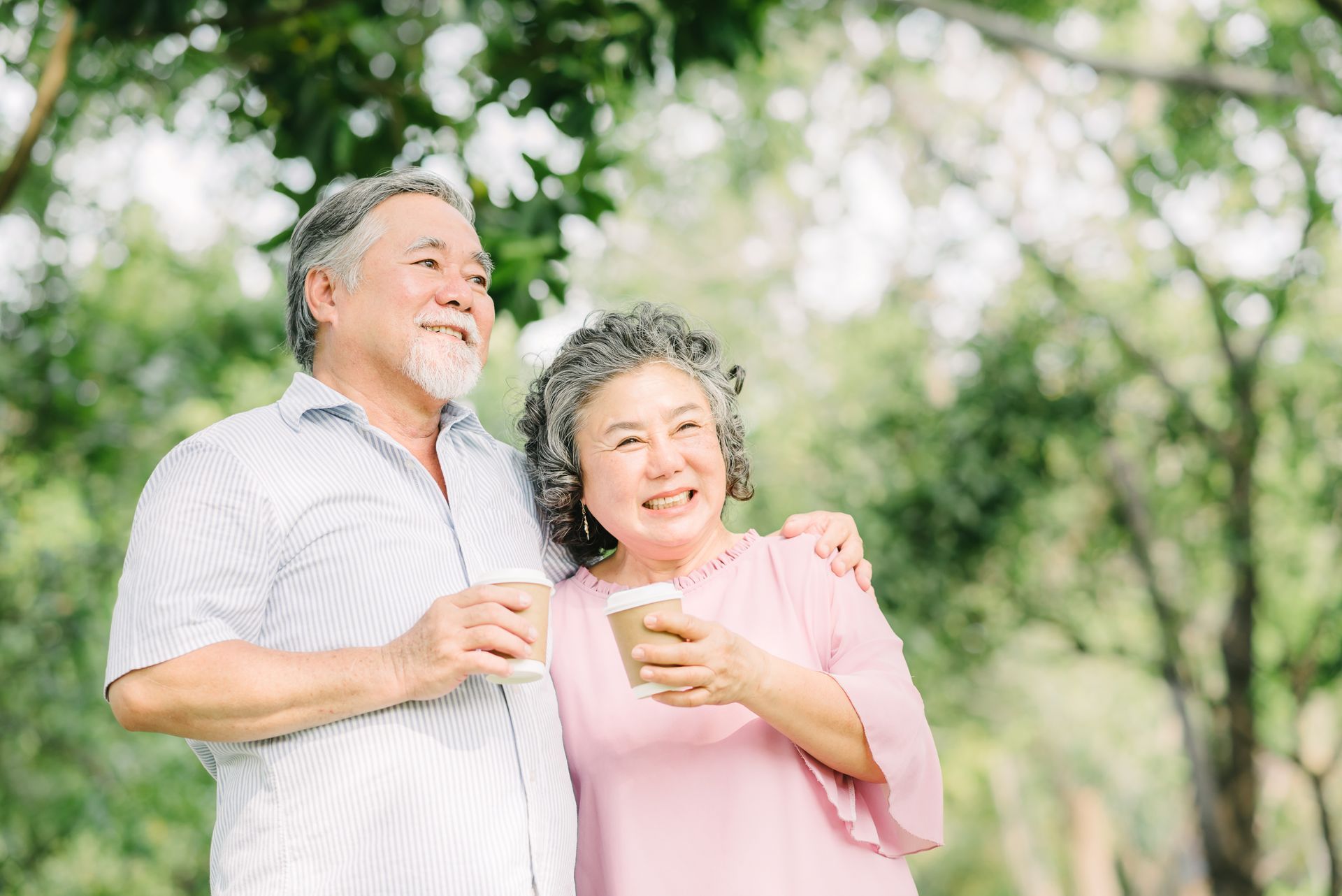 Retired couple standing in front of trees holding coffee mugs.