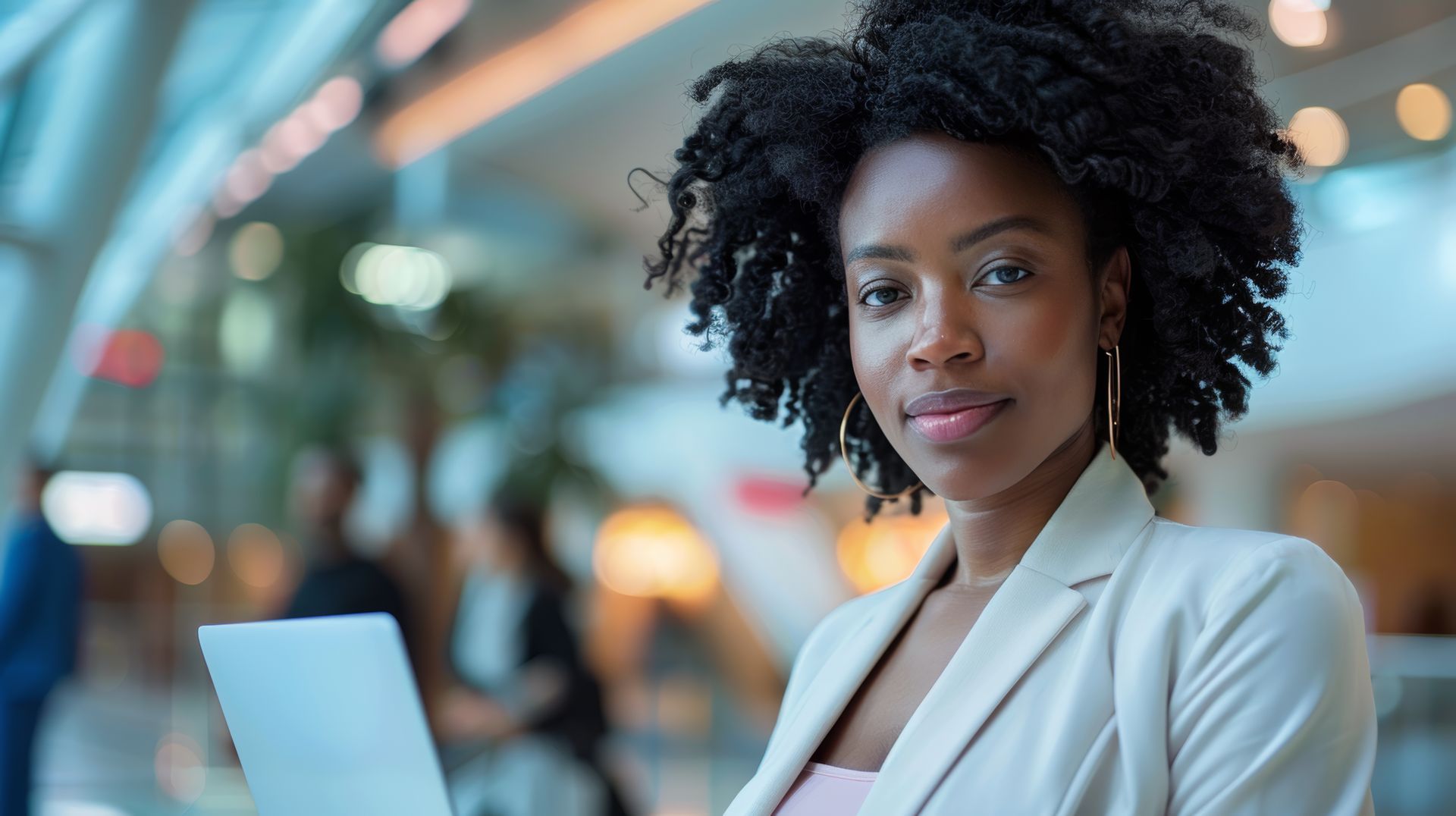 Dark skinned young adult woman in a white suit facing camera. Her lap top is open in front of her. 