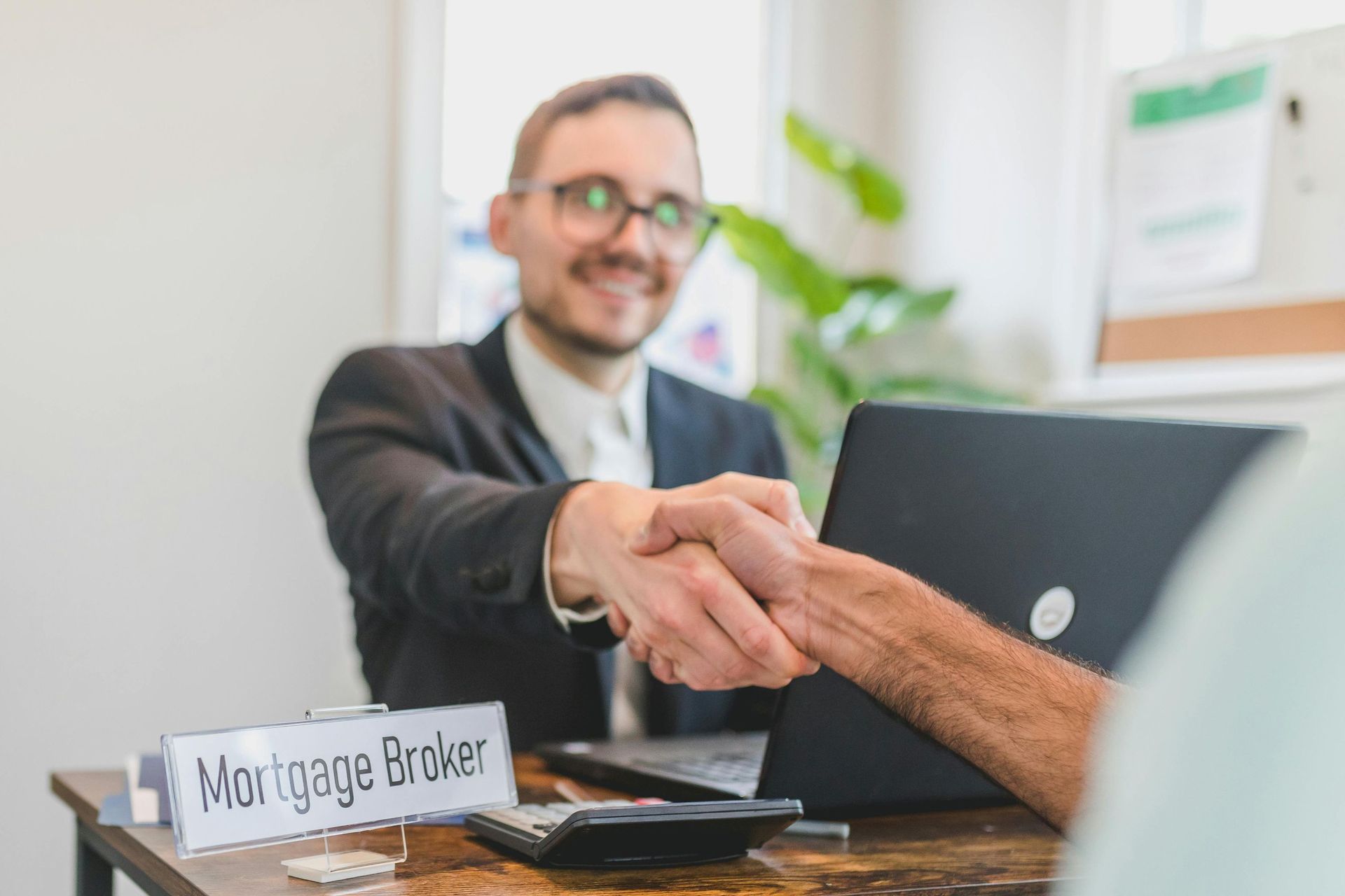 Mortgage broker shaking hands with a client at a desk; a laptop and calculator are visible.