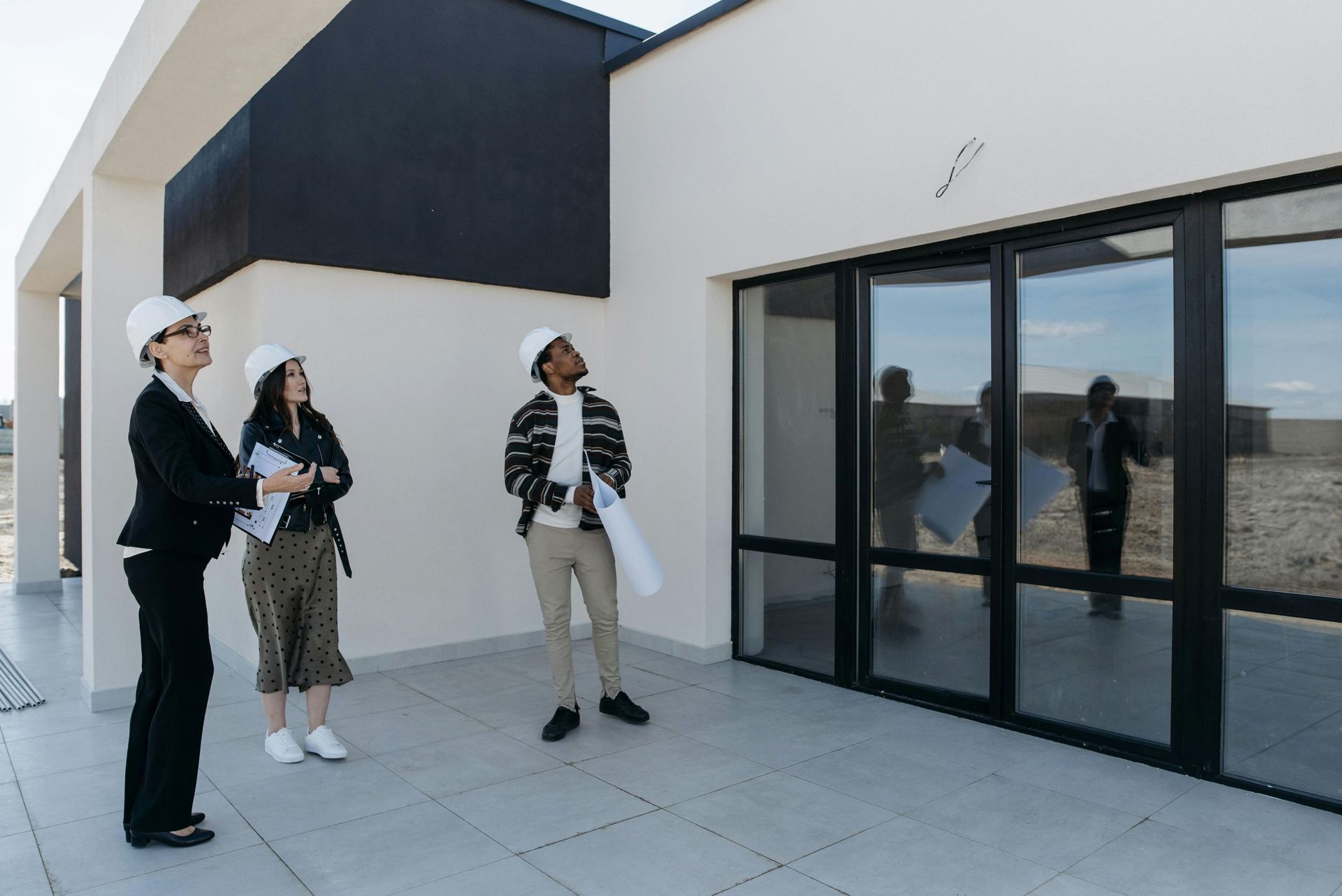 Three people wearing hard hats inspecting a modern building, holding blueprints.