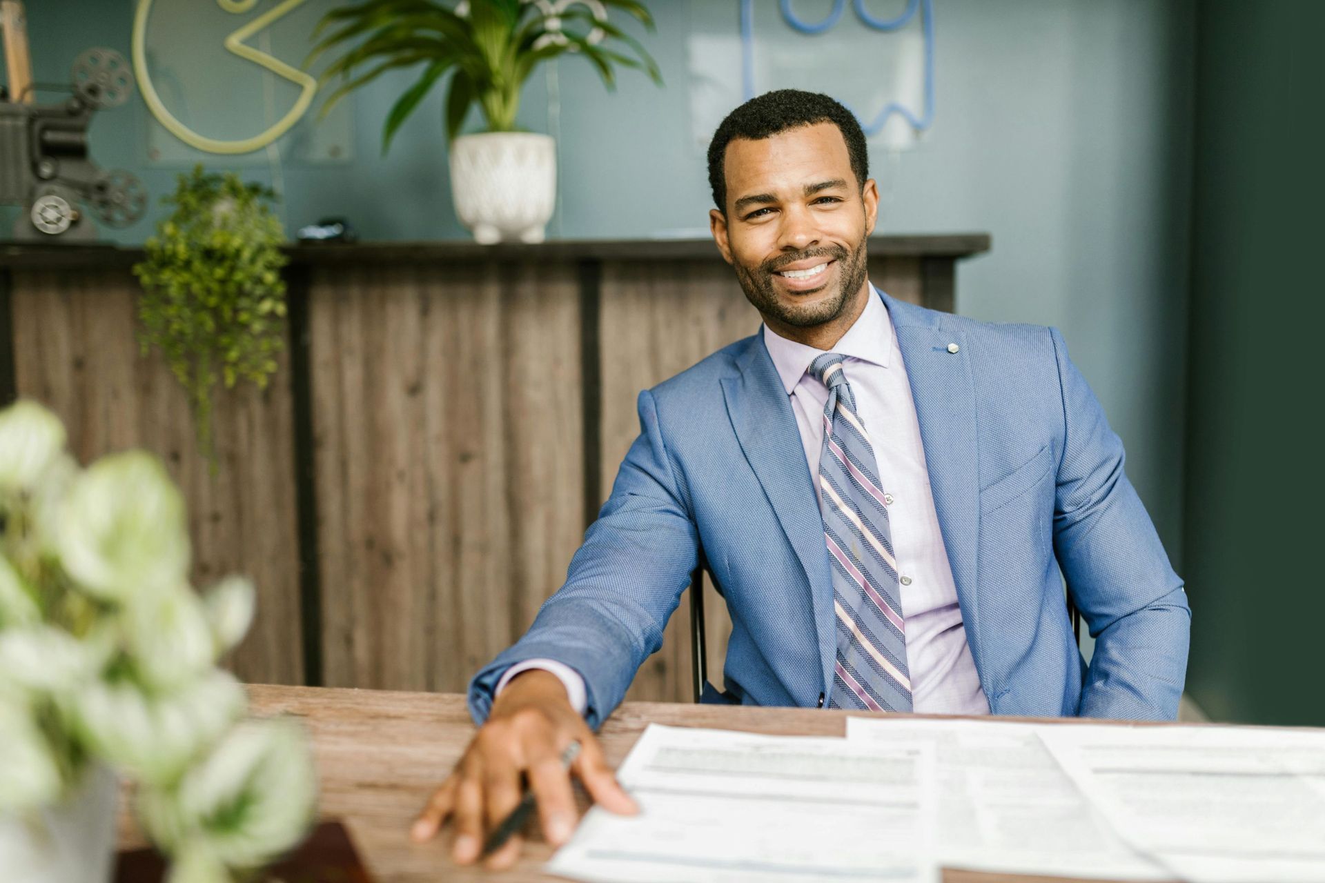 A smiling professional in a blue blazer and striped tie sits at a wooden desk with papers, in a modern office.