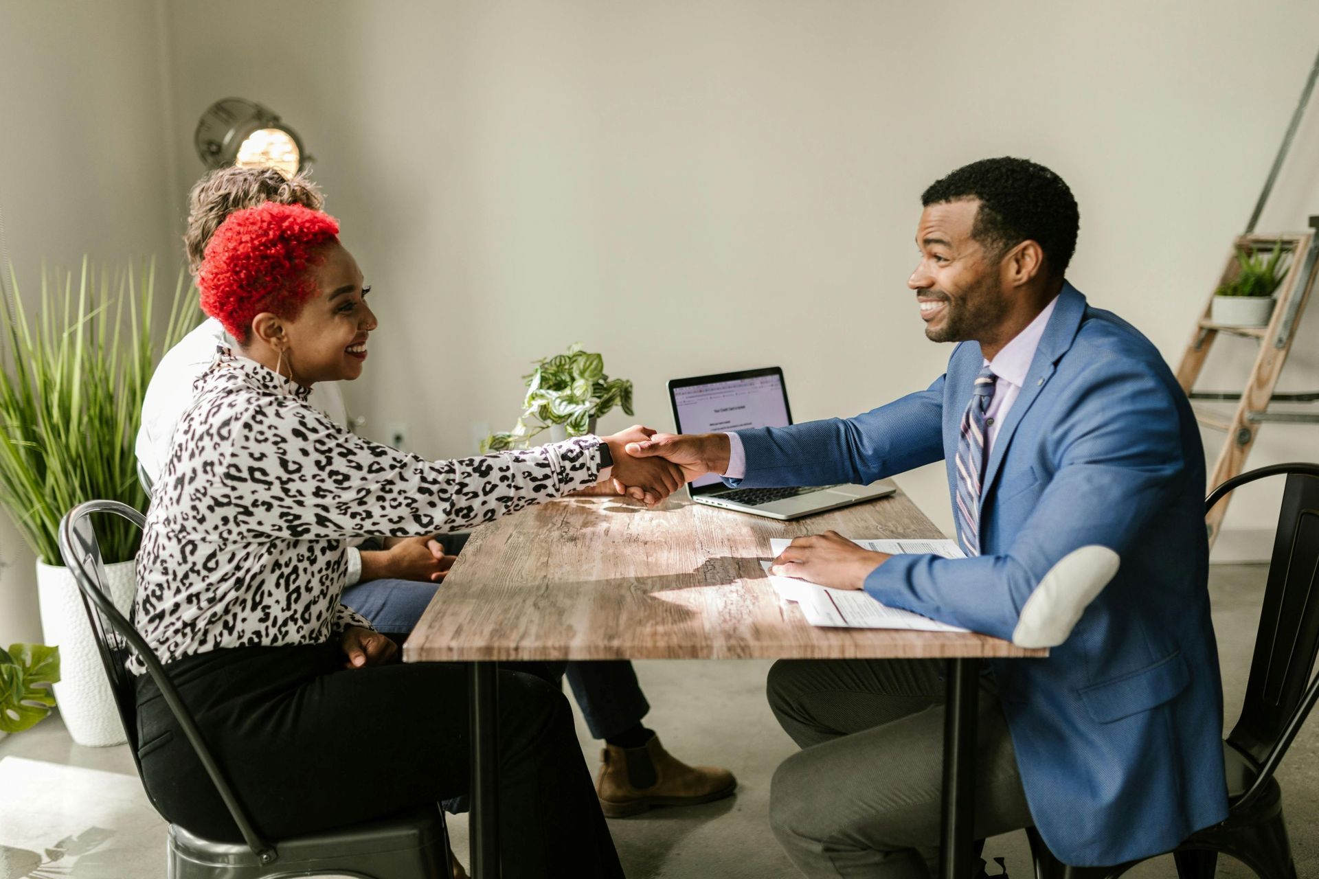 Woman with red hair shaking hands with a man in a blue suit at a wooden table; others are in the background.