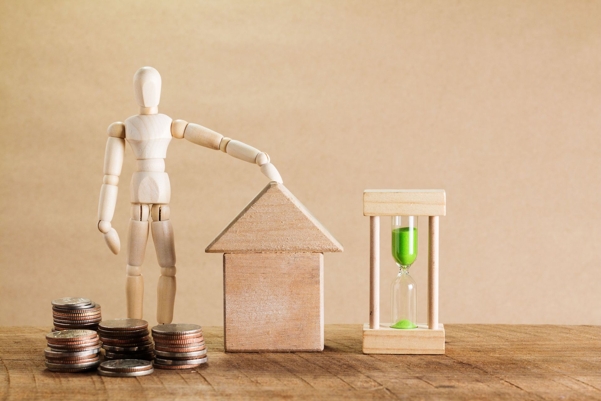 Wooden mannequin beside house model, coin stacks, and green hourglass on a table