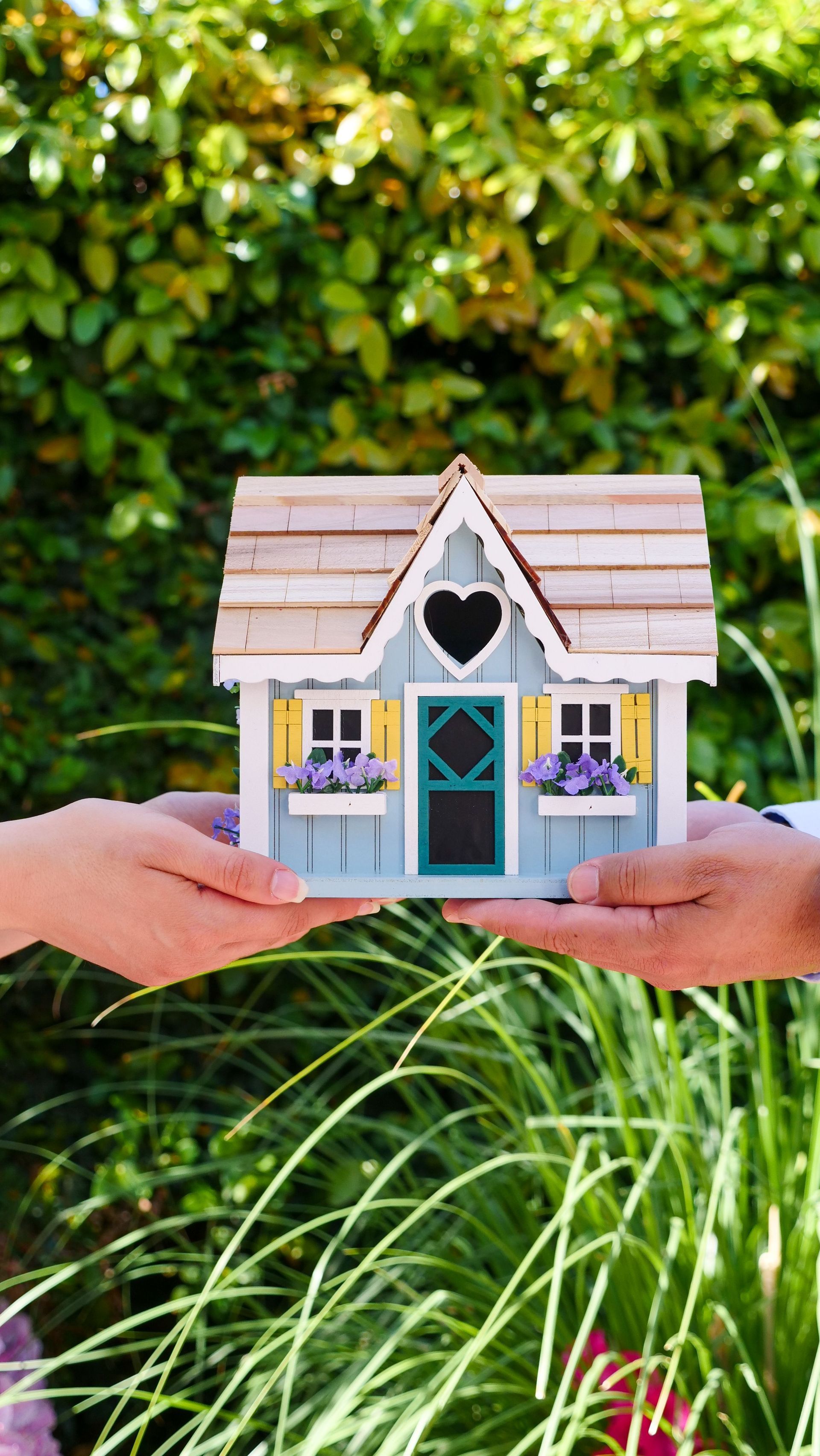 Hands holding a small pastel birdhouse in a green garden with flowers.